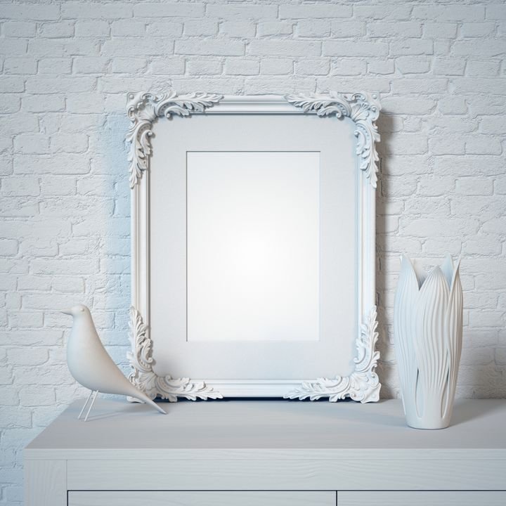 A white ornate frame rests on a light surface against a white brick wall, flanked by a bird figurine and a vase.